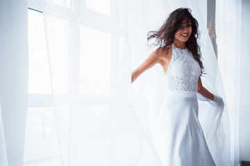 Shiny photo. Beautiful woman in white dress stands in white room with daylight through the windows