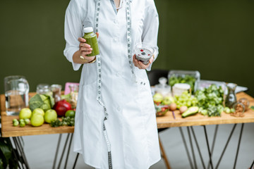 Nutritionist in medical gown holding salad and smoothie drink with healthy food on the background,...