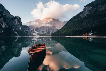 Clouds is on the top. Wooden boat on the crystal lake with majestic mountain behind. Reflection in the water. Chapel is on the right coast