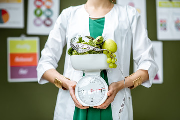 Young woman nutritionist in medical gown holding weights full of healthy products standing on the...