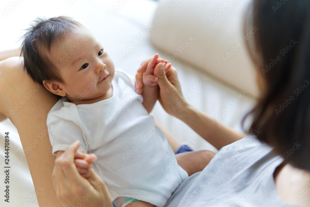 Wall mural asian newborn baby sitting on mother knees. she is looking at her mom and smile. mother holding baby