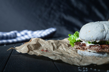 Burger with meat, sauce, salt and vegetables on craft paper. Snack with a blue bun on a dark wooden background with insta tint. Close up view on american fast-food. Copy free space for logo, text