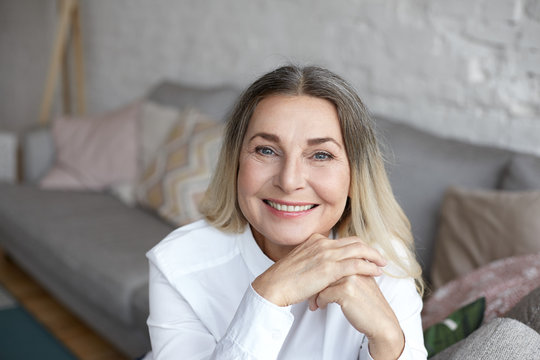 Picture of cheerful grandmother with blue eyes and shiny wrinkled skin spending day at home, sitting in living room with modern interior deisgn and smiling broadly at camera. People and lifestyle