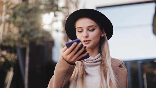 Business lady in black hat sending audio voice message on cellphone at outdoor talking to mobile assistant. Girl using smartphone voice recognition, dictates thoughts, voice dialing message