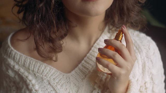Close up: young beautiful curly lady holding, using luxury perfume in orange glass bottle. Model wearing warm winter knitted sweater.