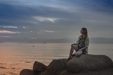 Fototapeta premium Girl on the rocks above sea and evening sky with sunset and blue clouds