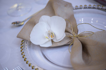 Obraz na płótnie Canvas Fine dining table setting featuring transparent plates, beige linen napkin with natural orchid and golden decorations and silverware in the order of use, ready for guests at a formal event or wedding