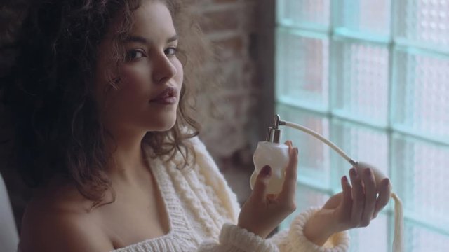 Close up: Young beautiful fashionable lady spraying, using, holding luxury perfume in vintage white glass bottle with pump. Model wearing warm winter knitted sweater