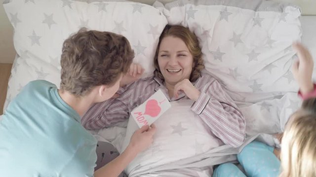 Mothers Day - Children surprise their Mum with presents in bed. Happy Family - beautiful Mother with her Son and Daughter. Girl and boy congratulates mom and gives her postcard.