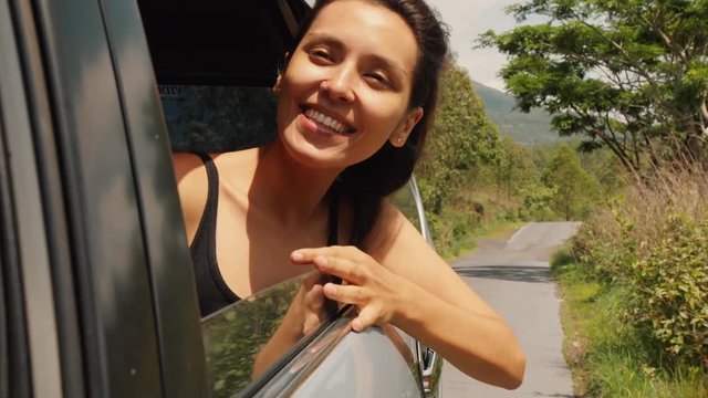 Happy Young Mixed Race Woman Leaning Out Side Car Window, Looking Around, Smiling and Enjoying Warm Summer Headwind. 4K Slowmotion. Bali, Indonesia.
