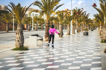 Fototapeta na wymiar Skater girl skating alone on the sidewalk at the edge of the beach with pink coconut trees and pink dressing. Playing sports with roller in line
