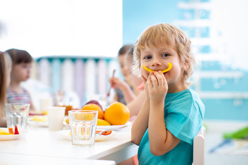 Funny kid boy eating fruits in kindergarten dining room. Child shows smile from paprika slice