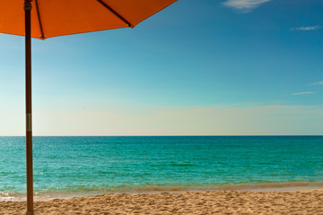 Orange beach umbrella on golden sand beach by the sea with emerald green sea water and blue sky and white clouds. Summer vacation on tropical paradise beach concept. Skyline between sea and sky.