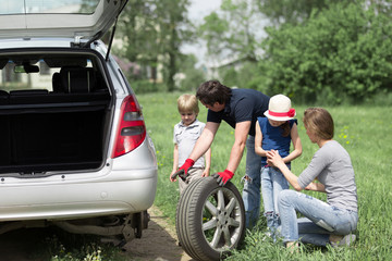 man replaces a damaged tire of a family car during a family trip