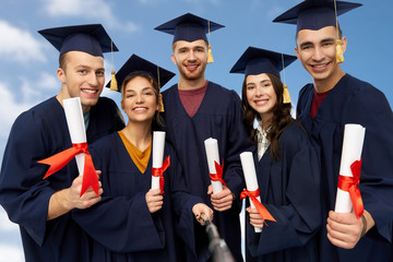 education, graduation and people concept - group of happy graduate students in mortar boards and bachelor gowns with diplomas taking picture by slfie stick over blue sky and clouds background