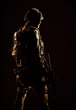 Low key studio shoot of army special forces soldier, commando fighter in mask, ballistic glasses, tactical helmet and battle uniform, holding short barrel service rifle, looking back over shoulder