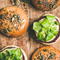 Flat-lay of healthy vegan burgers with quinoa beetroot patties, avocado cream and green sprouts over wooden background, top view, copy space, square crop. Vegetarian, alcaline diet food