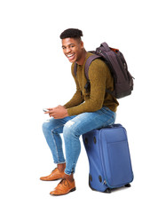 happy african american travel man sitting on suitcase with mobile phone against isolated white background