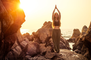 stylish redheaded chestnut blonde young girl in indie bohemian bo-ho style clothes black stylish top.boho woman long hair,makeup and green feathers in head practicing yoga asanas on the rocks by sea - obrazy, fototapety, plakaty