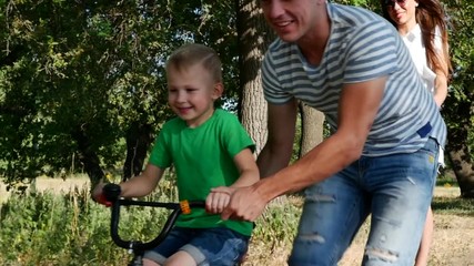 Happy family and carefree childhood. Happy family, mom, dad and son walking in nature. The father helps his son to accelerate on the bike. Stock footage. - Powered by Adobe