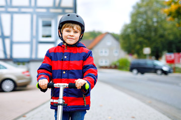 cute little school kid boy with helmet riding on scooter in park nature. children activities outdoor in winter, spring or autumn. funny happy child in colorful fashion clothes.