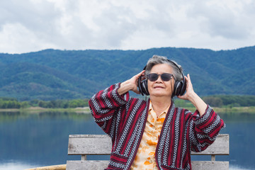 Portrait of happy senior woman listening music on headphone for relaxing with side the lake. Relaxing time concept.