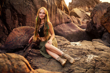 full-length portrait young and beautiful long-haired woman in a stylish skirt and top posing on the rocks beach .feathers dreadlocks in hair and makeup boho brown style dress fashion shoes boots
