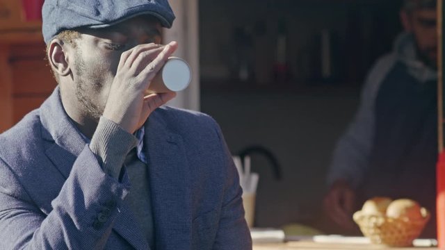 Up-section shot of young African American man drinking coffee near food booth and checks social media on tablet computer