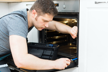 Professional handyman in overalls repairing domestic oven in the kitchen.