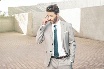 Young attractive serious businessman is standing next to a concrete wall background wearing light classic tuxedo. Urban working style.