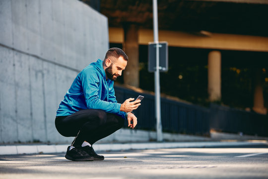 Young Caucasian bearded sporty man with short hair in sportswear crouching on the street and looking at smart phone. Healthy lifestyle concept.