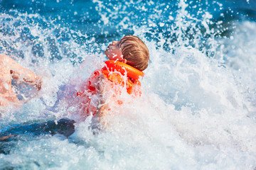 Little boy in orange life jacket swimming in the sea with waves with big splashes (selective DOF)