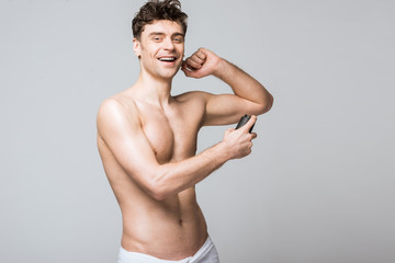 Fototapeta na wymiar cheerful shirtless man spraying deodorant, isolated on grey