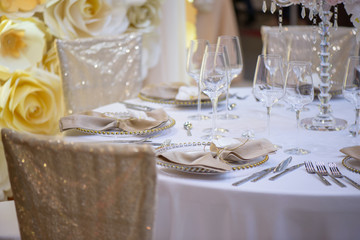 Sitting arrangement at a formal event or fine dining restaurant featuring transparent plates with golden details, glassware and silverware in the order of use set against a paper flowers wall