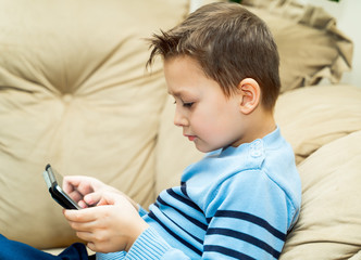 Side view of a nice boy sitting on the sofa and playing on the tablet. The picture of a casual little boy on the couch background. Close-up