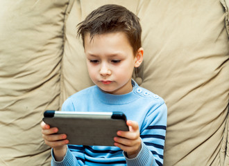 Serious boy watching something interesting from digital tablet sitting on a sofa in the living room at home. Gadgets and technology concept