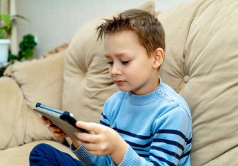 Little good-looking boy sitting on sofa and playing game on digital tablet. Portrait of a young child at home watching cartoon on the laptop.
