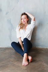 Naklejka premium Model blonde barefoot blue jeans white shirt sitting on floor against gray wall.