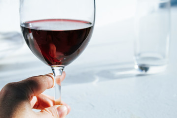 Woman's hand holding a glass of red wine in front of a white wall with bright summer light