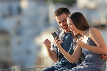 Excited couple celebrating online phone news outdoors