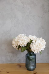  A bouquet of white hydrangea flowers in a vase on a wooden floor near a gray wall. Hello spring, March, April, May