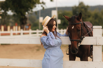 Beautiful pregnant woman stand near the horse. Lady in knitted hat and blue dress. Beautiful farm animal. Brown Hors