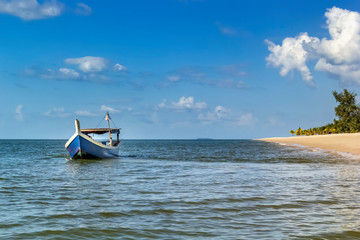 Traditional asian wooden boat on the waves near the sandy beach with palm trees against the blue sky with clouds on a sunny day