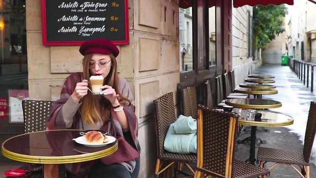 Beautiful girl with long hair, red lipstick, glasses, beret sitting having breakfast at the french cafe, drink coffee with croissant and smiling