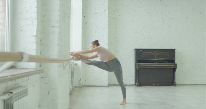 Elegant pretty ballerina with perfect body doing stretching leg and spine exercises at ballet barre in dance studio before rehearsal. Graceful female dancer in sport outfit warming up before dancing.