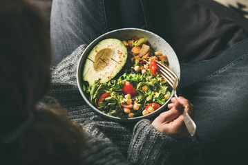 Healthy vegetarian dinner. Woman in jeans and woolen sweater holding bowl with fresh salad, avocado...