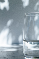 Close up of a glass of water standing in front of a white wall with shadows of leaves on a bright, sunny day