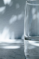 Close up of a glass of water standing in front of a white wall with shadows of leaves on a bright, sunny day