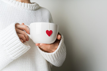 Female in knitted woolen light sweater holding white mug in hands with heart of teabag. Valentine's day mockup background with love and copy space