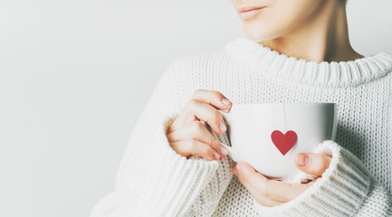 Beautiful woman in warm knitted woolen light sweater holding white mug in hands with heart of teabag with love and copy space. Concept for Valentine's day mockup.